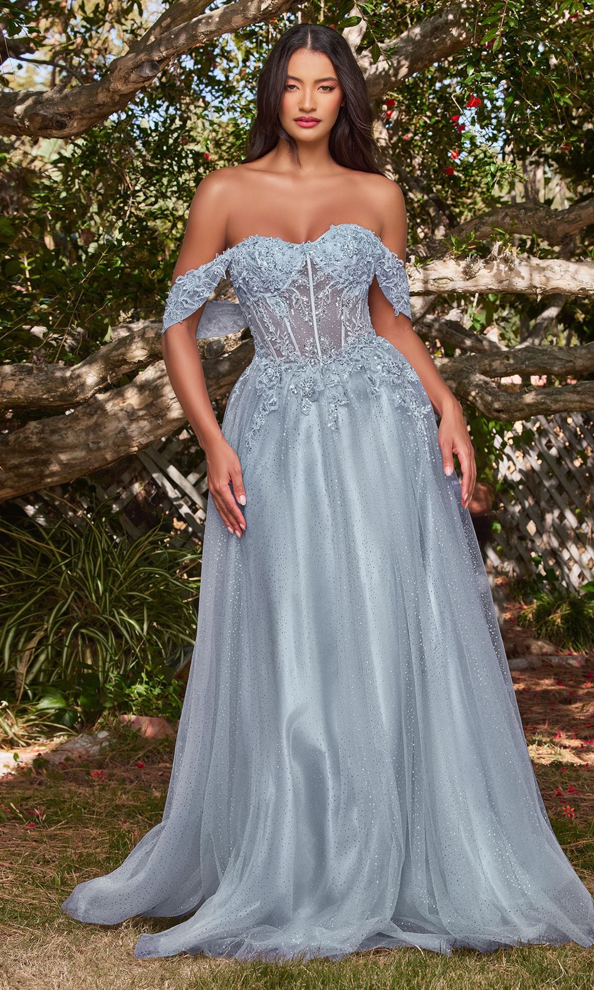 Woman wearing the Off-Shoulder Long Glitter A-Line Prom Dress CD0198 in light blue, featuring embroidered lace and flowing tulle, stands outdoors surrounded by lush trees and greenery.